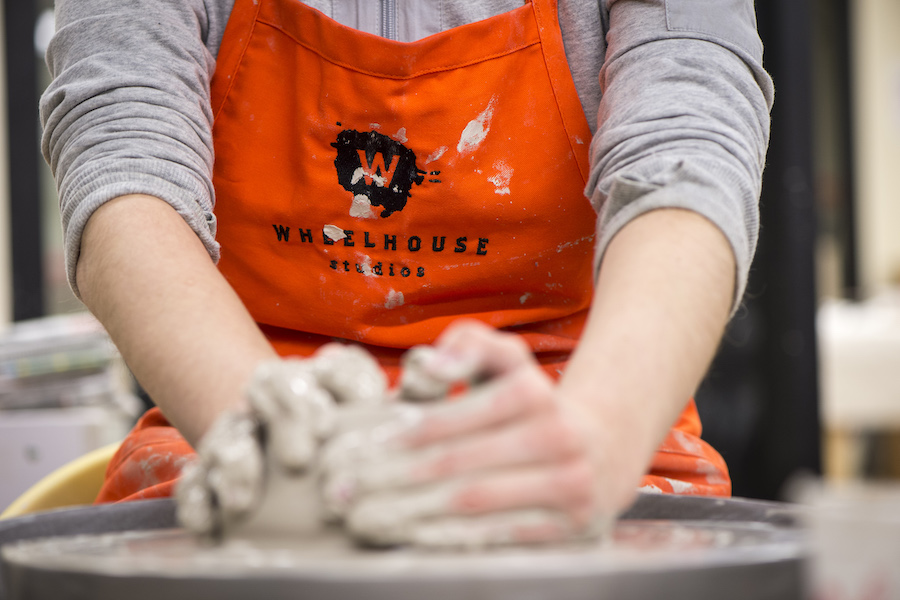 Close up of a person throwing a pot on a pottery wheel, wearing an orange apron with Wheelhouse Studios logo